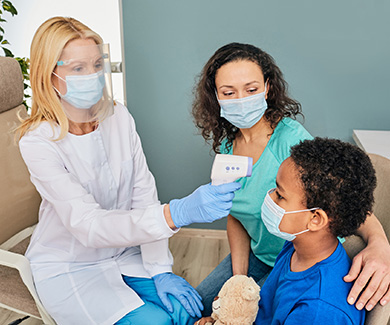 A masked medical assistant takes the forehead temperature of a child.