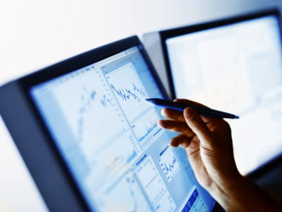 A hand holding a pen pointing to graphs on a computer screen.