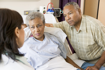 A photograph shows a woman in a hospital bed and a man standing at the bedside, holding her hand. They are listening to a health professional, who is standing with her back to the camera.